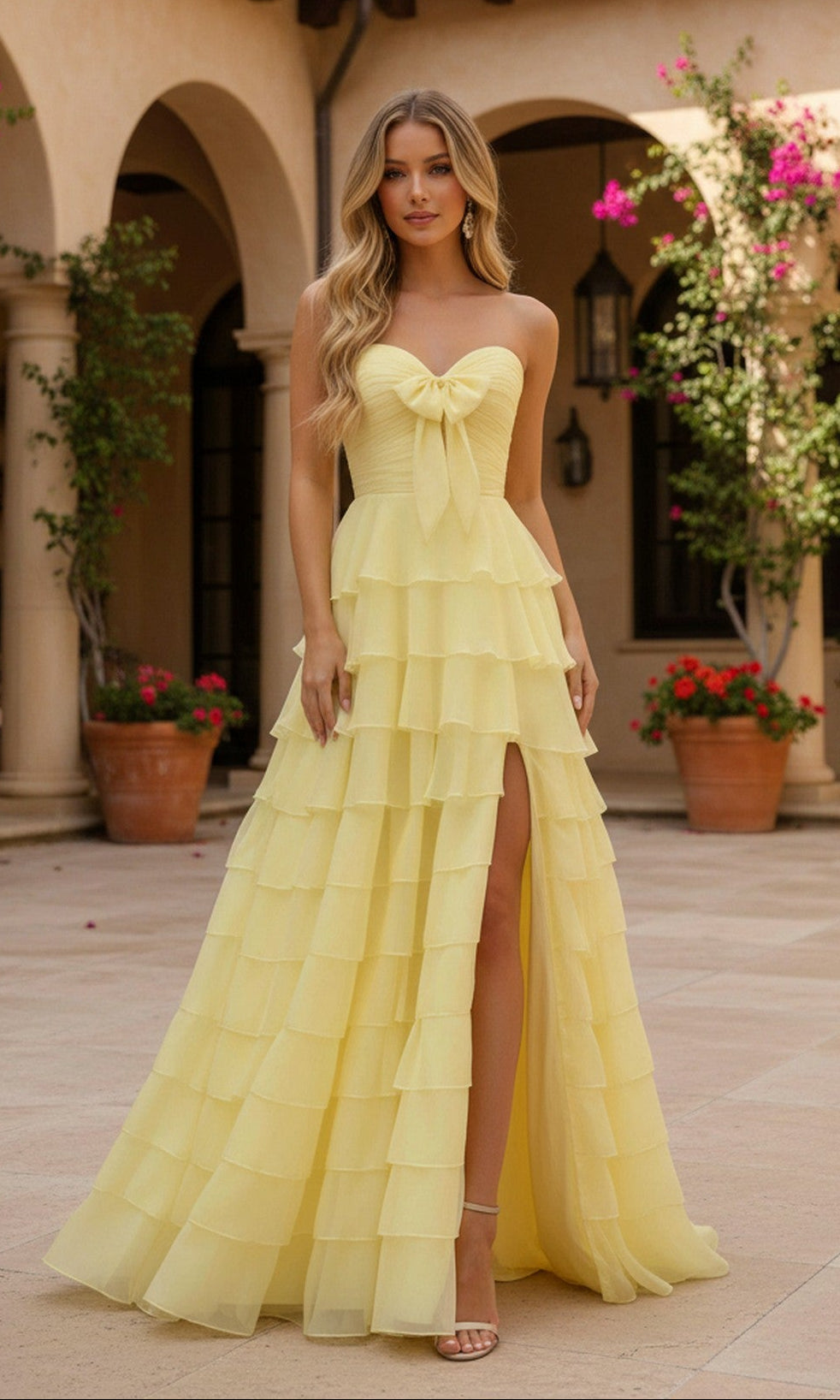 A woman wears the PromGirl Exclusive 250233B, a light yellow strapless prom dress with a tiered ruffle skirt and a bow-accented bodice, standing in a courtyard with potted plants and arched architecture.
